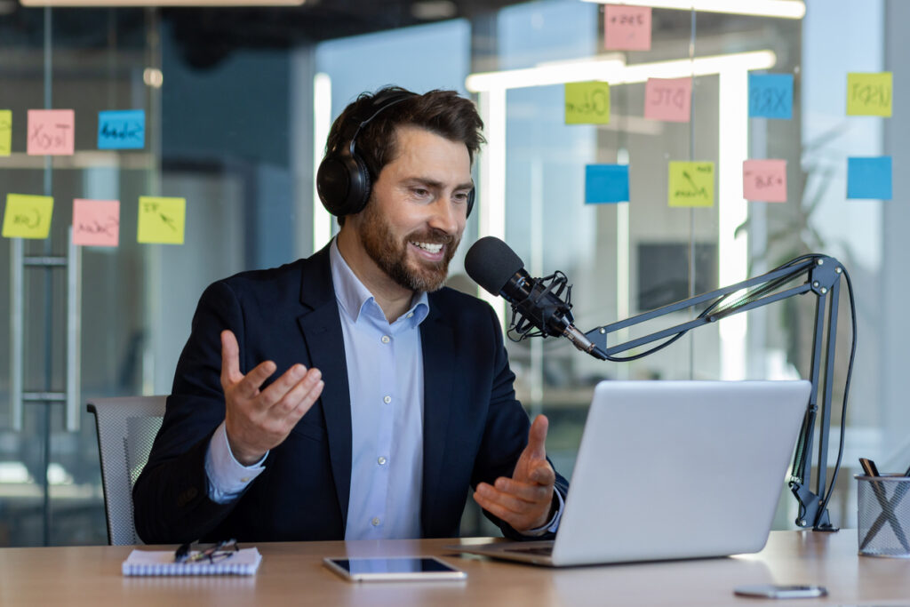 Man recording podcast in office with microphone, headphones, laptop, expressing enthusiasm and professionalism.